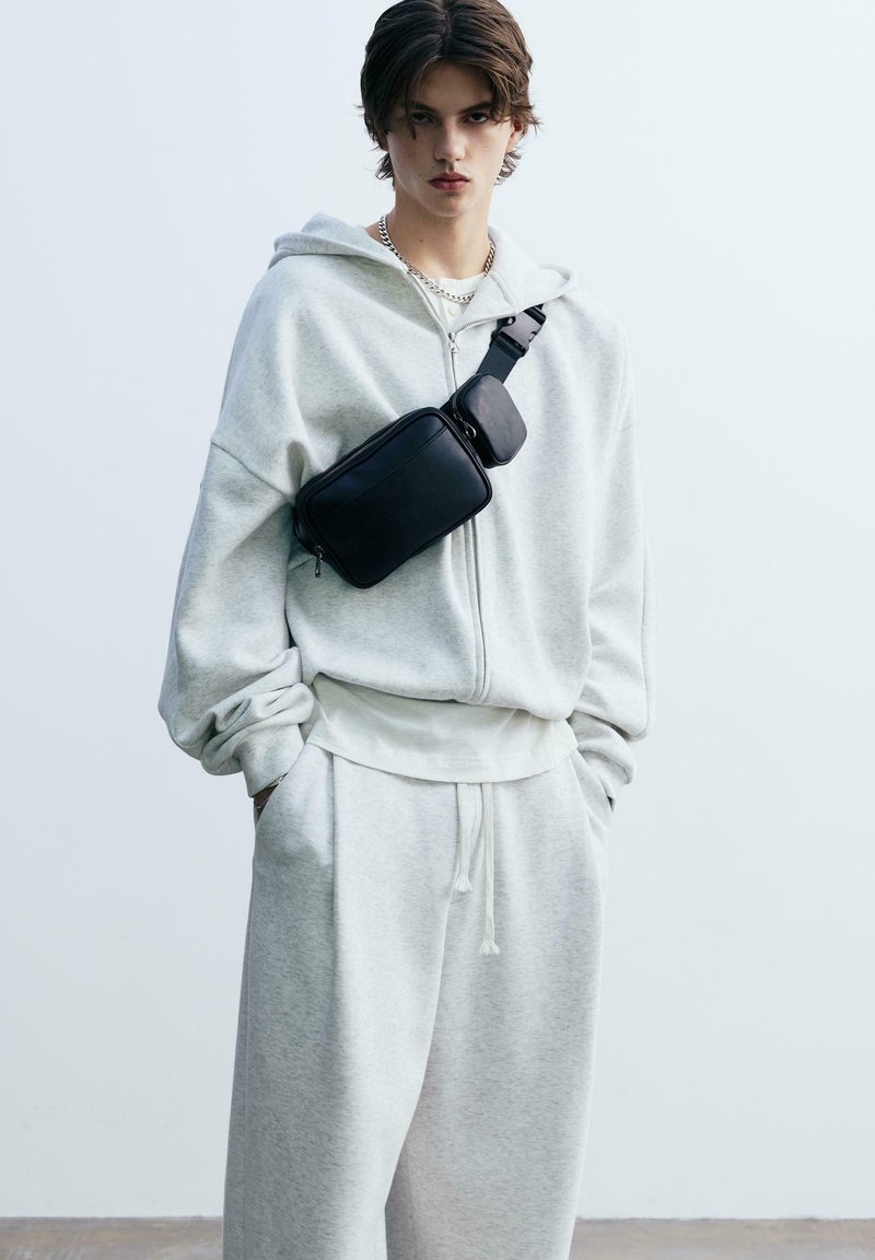 Grå, strukturerad huvtröja med dragkedja och framfickor, tillsammans med matchande sweatpants. En svart axelväska bärs över bröstet.