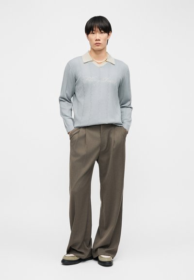 Νεαρός άνδρας που στέκεται, φορώντας ένα ανοικτό γαλάζιο πλεκτό πουλόβερ με γιακά και κείμενο, καφέ παντελόνια με φαρδιά πόδια, περιδέραιο με πέρλες και ανοιχτά αθλητικά παπούτσια.