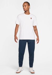 Vit bomulls t-shirt med en liten flerfärgad logotyp; matchad med marinblå träningsoverallbyxor; bärs med krämfärgade sneakers med bruna accenter.