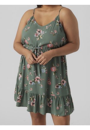 Kobieta w zielonej sukience w kwiaty na cienkich ramiączkach z wiązaniem w pasie, ręce złożone przed sobą, na jednolitym tle.