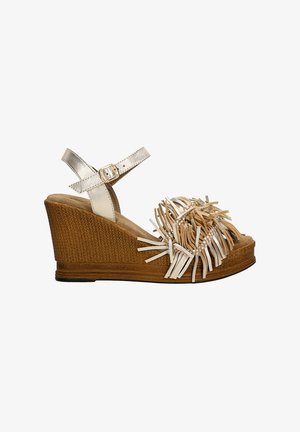 Klínová sandála s hnědou tkanou bází, metalicky stříbrným náramkem kolem kotníku a dekorativním třásním z barevných pásků na oblasti prstů.