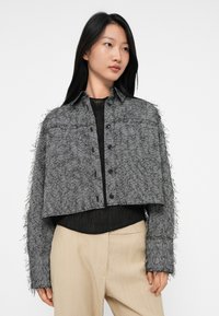 Vrouw in een cropped grijze herringbone textuurjas over een zwarte top en beige broek, die iets naar rechts kijkt tegen een witte achtergrond.