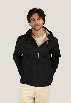 Homme portant une veste à capuche noire zippée avec des accents orange sur une chemise claire et un pantalon beige, debout les mains derrière le dos.