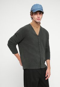 Sötétzöld kötött kardigán V-nyakkivágással, gombos záródással és hosszú ujjakkal. Bézs garbó fölött viselve, farmer sapkával párosítva.