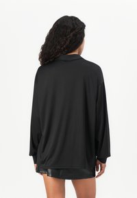 Žena s dlhými kučeravými vlasmi má na sebe voľnú čiernu košeľu s dlhým rukávom a krátku čiernu koženú sukňu, otočená chrbtom, na bielom pozadí.