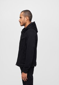 Giacca di jeans nera con cappuccio foderato in pile, caratterizzata da una chiusura con bottoni, tasche laterali e un tessuto leggermente strutturato. Vista laterale.