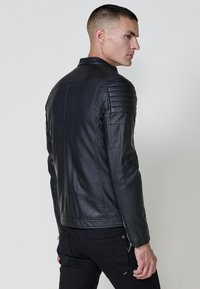 Homme en veste en cuir matelassé noir et pantalon noir, se tenant de profil contre un fond gris uni.