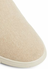 Béžová plátěná bota s texturovaným svrškem a zaoblenou špičkou, která má světlou gumovou podrážku a bezšvový design.