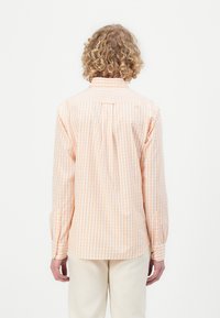 Camicia a quadri arancione e bianca con maniche lunghe, colletto alla francese e dettagli con piega laterale sul retro. Indossata con pantaloni color crema.