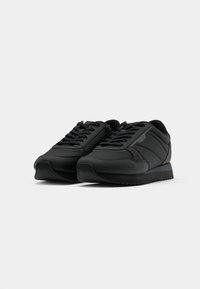 Paire de baskets basses noires avec lacets et fermetures éclair latérales, présentée sur un fond uni gris clair.