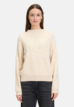 Helle beige Strickjacke mit einem strukturierten Blumenmuster, rundem Ausschnitt und langen Ärmeln, kombiniert mit dunklen Jeans mit Nietenakzenten.