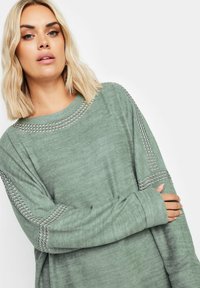 Ljusgrön långärmad topp med en mjuk textur, prydd med metalliska nitar längs axlarna och ärmarna. Rund halsringning.