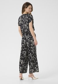 Svart byxdress med vitt blommönster, korta ärmar, lös passform, cinchad midja, vida ben och platta sandaler. Slät tygstruktur.