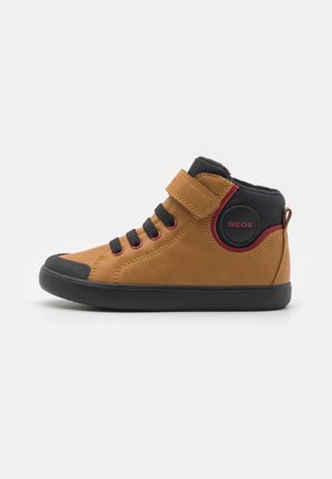 Zapato alto marrón con tira de velcro, acentos negros, punta de goma y un logo circular de GEOX en el lado. Parte superior de ante texturizado.