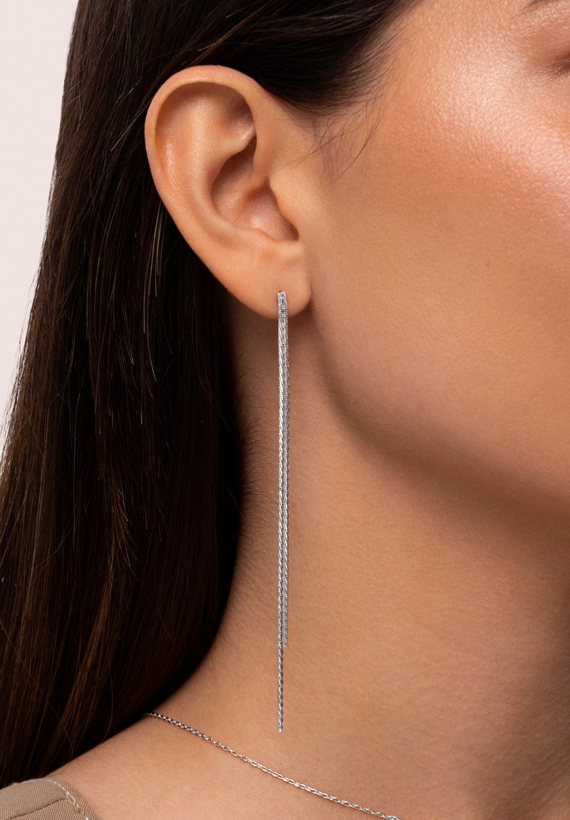 Silver dangling earrings with a sleek, elongated design. Textured finish and minimalist aesthetic. Worn on the left ear with a subtle chain necklace.