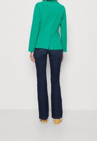 Veste verte à manches longues avec une fente dans le dos, assortie à un jean flare en denim foncé et des sabots en bois. Design simple, texture lisse.