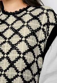 Femme portant un gilet en tricot noir et blanc avec un motif en losange sur une chemise à manches longues blanche avec un col en dentelle.