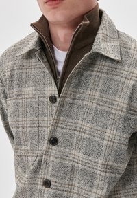 Jachetă gri cu carouri, cu accente maronii; material texturat, guler ascuțit, fermoar frontal și trei nasturi negri.
