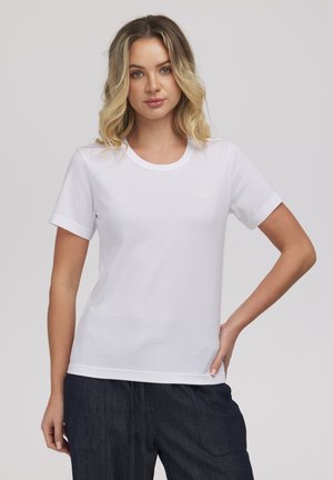 Młoda kobieta z blond, falującymi włosami, ubrana w prosty biały T-shirt z krótkim rękawem i ciemne dżinsy, stoi z jedną ręką na biodrze.