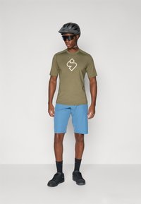 Oliivinvihreä lyhythihainen paita, jossa on beige logo, yhdistettynä vaaleansinisiin shortseihin. Asusteina musta kypärä ja aurinkolasit. Mustat kengät täydentävät asua.