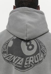 Grijze hooded sweatshirt met een grote zwarte afbeelding van een biljartbal en de tekst "Santa Cruz" op de achterkant, gemaakt van een zachte, textuurrijke stof.