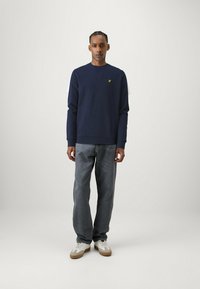 Sweatshirt blu navy concollo rotondo e polsini a costine, abbinato a jeans grigi e sneakers bianche, con un piccolo logo verde sul petto.