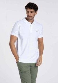 Chemise polo blanche avec col classique, manches courtes et un petit logo brodé sur la poitrine. Le tissu semble lisse et léger.