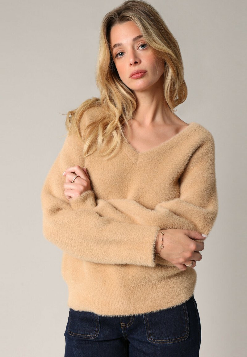 Beige, donzige trui met een v-hals, lange mouwen en een losse pasvorm. Zachte textuur en minimalistisch ontwerp. Donkere jeans complementeren de look.