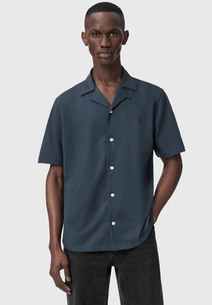 Mand iført en mørkeblå kortærmet skjorte med knapper og sorte jeans, stående med den ene hånd i lommen mod en ensfarvet baggrund.