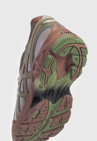 Scarpa sportiva con suola in gomma marrone e verde texturizzata. La parte superiore presenta pannelli in rete e accenti sintetici in tonalità variate.