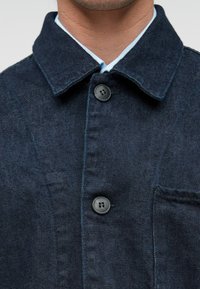 Chaqueta de denim oscuro con cuello abatible, dos botones grandes y un bolsillo en el pecho. La tela tiene una textura sutil y un color azul profundo.