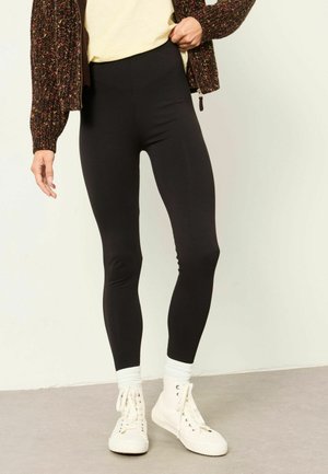 Persona con mallas negras, calcetines blancos, zapatillas altas blancas, un cárdigan de punto multicolor y una camisa de color claro.