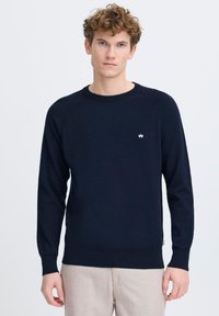 Suéter azul marino hecho de suave tejido de punto, con un clásico escote redondo y un pequeño logo blanco en el pecho. Mangas largas y puños de canalé.