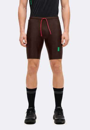 Homme portant un short marron moulant avec un cordon rose, des chaussettes noires mi-mollet avec des rayures grises, et des chaussures de course noires, debout.