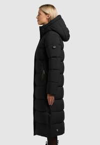 Long manteau matelassé noir avec capuche, présentant un design matelassé, des poches zippées et un patch logo sur la manche. Texture lisse.