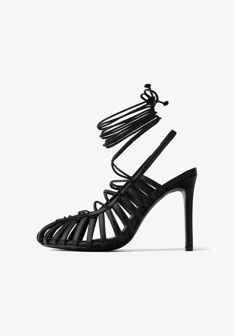 Sandal nera con tacco alto, dal design con cinturini e punta aperta, caratterizzata da bande in pelle verticali e una sottile cinghia alla caviglia. Finitura lucida.