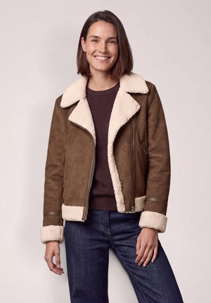 Femme souriante porte une veste marron avec bordure en peau lainée crème, associée à un pull foncé et un jean bleu sur un fond uni.