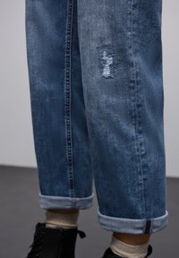 Jeans de denim en azul oscuro con parches desgastados, dobladillos enrollados y una textura desvaída. Presentan costuras en contraste y un corte recto.
