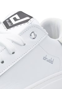 White sneaker featuring a smooth leather upper, white laces, a grey logo patch, and a distinctive circular hardware accent.