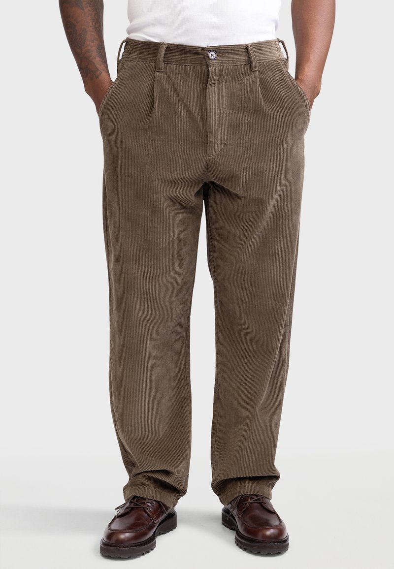 Pantalon en velours côtelé marron avec des rayures verticales, coupe droite, deux poches avant et fermeture boutonnée. Porté avec des chaussures marron foncé.
