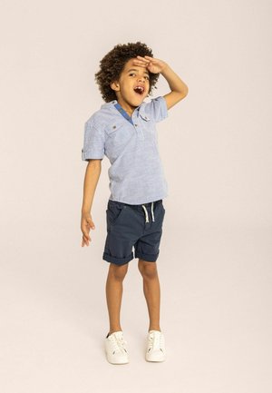 Maglietta a maniche corte a righe blu con colletto abbottonato, abbinata a pantaloni corti blu scuro e sneaker bianche. Il bambino sta posando con una mano sollevata.