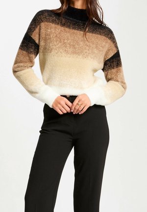Femme portant un pull flou dégradé noir, marron, beige et blanc avec un pantalon noir, debout les mains jointes à la taille.