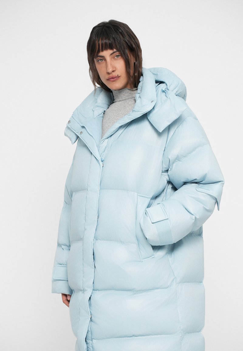 Doudoune bleu clair avec capuche, dotée d'une fermeture éclair, de poignets élastiques et d'un design matelassé. Texture lisse et brillante.