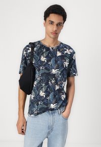 Tricou bleumarin cu flori, având crini albi și frunze verzi, confecționat dintr-un material moale; purtat cu blugi albaștri deschis și o geantă crossbody neagră.