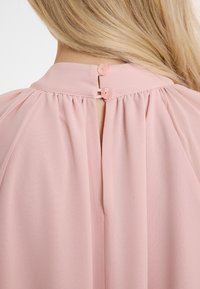 Hellrosa Bluse mit Knopfverschluss auf der Rückseite, plissierten Details und einem dezenten Ausschnitt am Hals. Weicher, fließender Stoff.