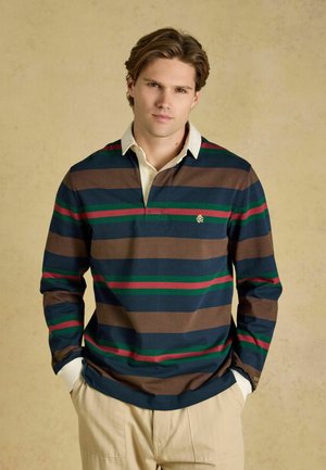 Jonge man die een gestreept rugbyshirt draagt met bruine, marineblauwe, groene en rode strepen, staand met handen in de zakken van een beige broek tegen een effen achtergrond.