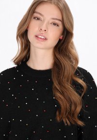 Fekete pulóver színes gyöngy díszítéssel, kerek nyakkivágással és puha textúrával. A modellen hosszú, hullámos haj és egyszerű fülbevalók vannak.