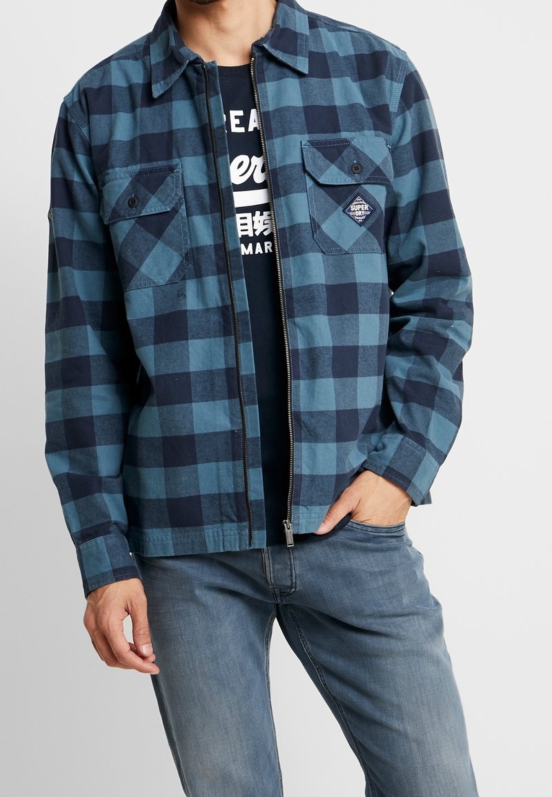 Giacca a quadri blu e navy con zip frontale, due tasche sul petto e patch con logo; indossata sopra una camicia scura e jeans grigi.