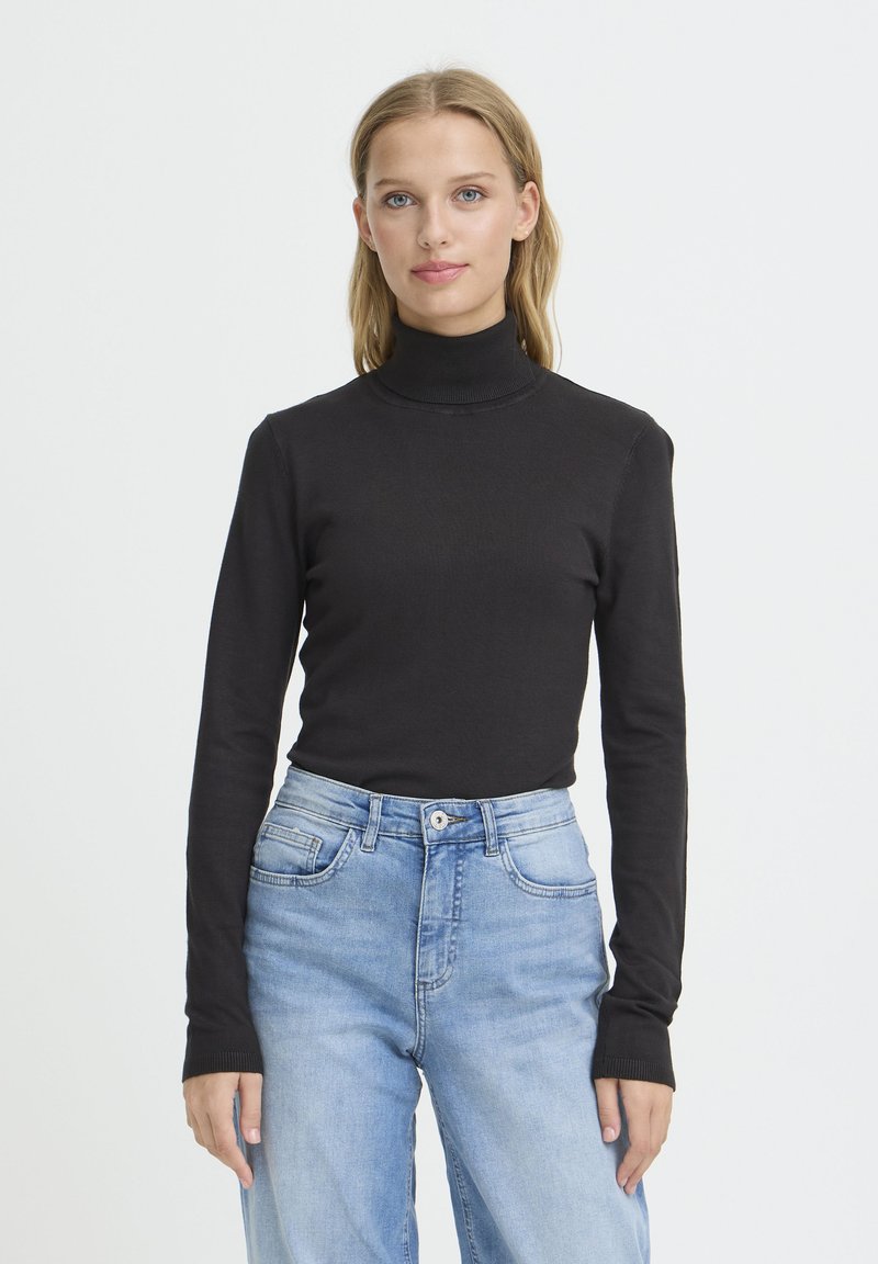 Femme aux cheveux blonds portant un pull à col roulé noir et un jean bleu clair, debout devant un fond blanc uni.