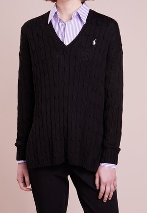 Persona con un suéter de punto negro con cuello en V sobre una camisa de cuello con rayas moradas claras y pantalones negros.